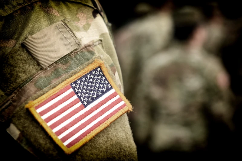 Close-up of a U.S. flag patch on the sleeve of a camouflage military uniform, with other blurred soldiers in similar uniforms in the background.